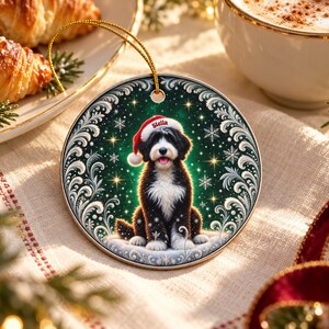 May include: A round ceramic Christmas ornament featuring a black and white dog wearing a red Santa hat with the name Stella printed on the brim. The dog is centered against a dark green background with white snowflakes and gold stars.