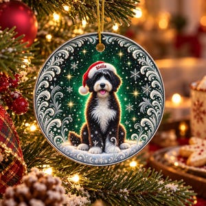 May include: A round ceramic Christmas ornament featuring a black and white dog wearing a red Santa hat with the name Stella printed on the brim. The dog sits against a dark green background with white swirling filigree and snowflake accents.
