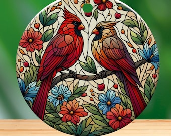 May include: Round ceramic ornament featuring two cardinals perched on a branch, surrounded by colorful flowers and foliage. The birds are depicted in shades of red, orange, and brown, with a stained-glass effect. A gold string is attached for hanging.