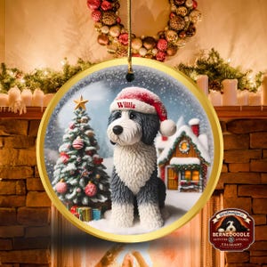 May include: A circular Christmas decoration depicting a Bernerdoodle dog wearing a Santa hat with the name "Willis". The ornament shows a winter scene with a Christmas tree and a snow-covered house. The decoration is trimmed in gold.