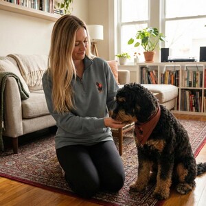 Pu&ograve; includere: Una felpa grigia con zip a un quarto di collo con un piccolo disegno di cane ricamato. Il cane &egrave; nero e marrone. Il cane indossa una bandana rossa. La felpa &egrave; abbinata a pantaloni neri. Un cane nero e marrone &egrave; seduto su un tappeto.