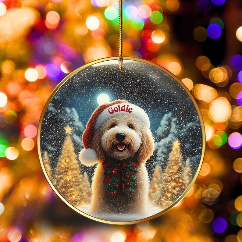 May include: Round Christmas ornament featuring a smiling dog wearing a Santa hat with the name "Goldie" and a festive garland. The background shows a snowy scene with Christmas trees and a full moon. The ornament has a gold-colored trim.