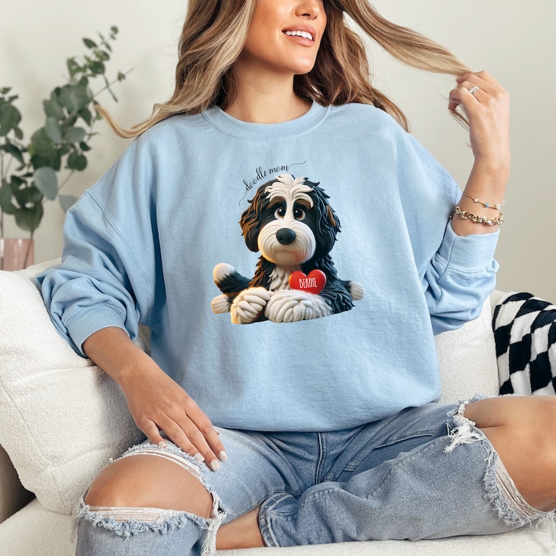 Unisex crewneck sweatshirt featuring an adorable claymation-style Bernedoodle dog illustration on the front, holding a red heart tag that can be personalized with a pet’s name. The back of the sweatshirt shows a cute rear-view Bernedoodle lying flat with detailed fluffy fur and oversized pink-paw pads, creating a playful “doodle mom” design. Ideal for Bernedoodle lovers, dog moms. Great for holidays, Mother’s Day, personalized pet gifts, and everyday cozy wear.