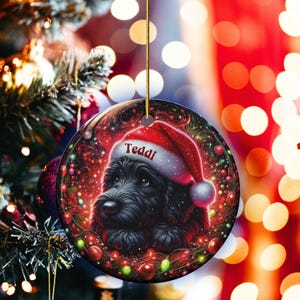 May include: Round Christmas ornament featuring a black dog wearing a Santa hat with the name "Teddi" on it. The ornament has a festive design with red, green, and gold accents, including Christmas lights and ornaments. It hangs from a gold string.