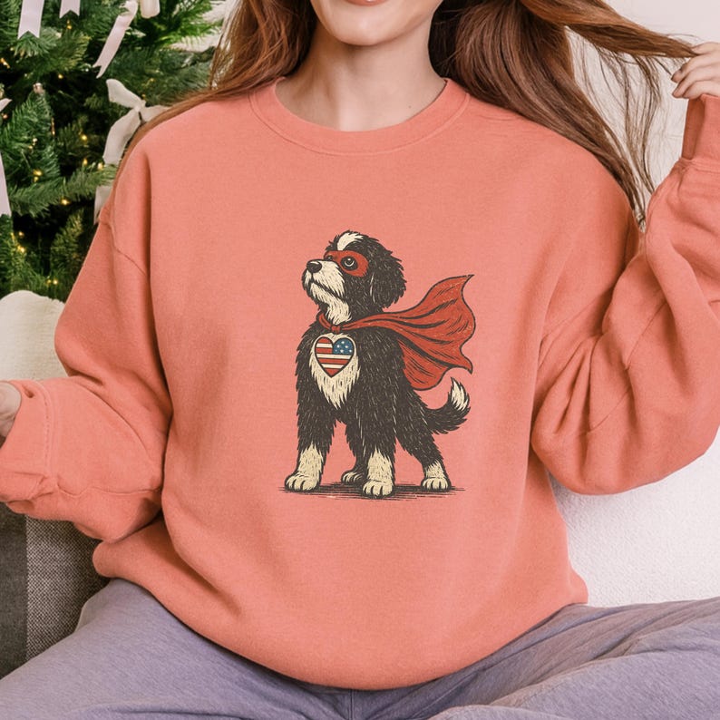 May include: A coral-colored sweatshirt featuring a cartoon dog dressed as a superhero. The dog has a red cape, a mask, and a heart-shaped emblem with an American flag design on its chest. The dog is standing.