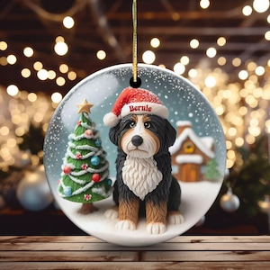 May include: A round Christmas ornament featuring a Bernese Mountain Dog wearing a Santa hat with the name "Bernie". The ornament has a Christmas tree and a small house in a snowy scene. The ornament is hanging from a gold string.