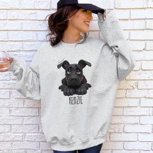 Puede incluir: Sudadera gris claro con una ilustraci&oacute;n de un perro negro y el nombre "KENZIE" en una fuente serif. El perro tiene un collar rojo. La sudadera tiene mangas largas y cuello redondo. Las palabras "dog mom" est&aacute;n escritas encima del perro.