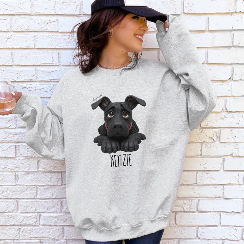 Puede incluir: Sudadera gris claro con una ilustraci&oacute;n de un perro negro y el nombre "KENZIE" en una fuente serif. El perro tiene un collar rojo. La sudadera tiene mangas largas y cuello redondo. Las palabras "dog mom" est&aacute;n escritas encima del perro.