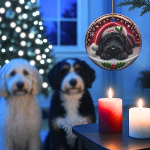 May include: A round Christmas ornament featuring a black dog wearing a Santa hat with the name "Teddi". The ornament is hanging in front of a Christmas tree and two lit candles. Two dogs are in the background.