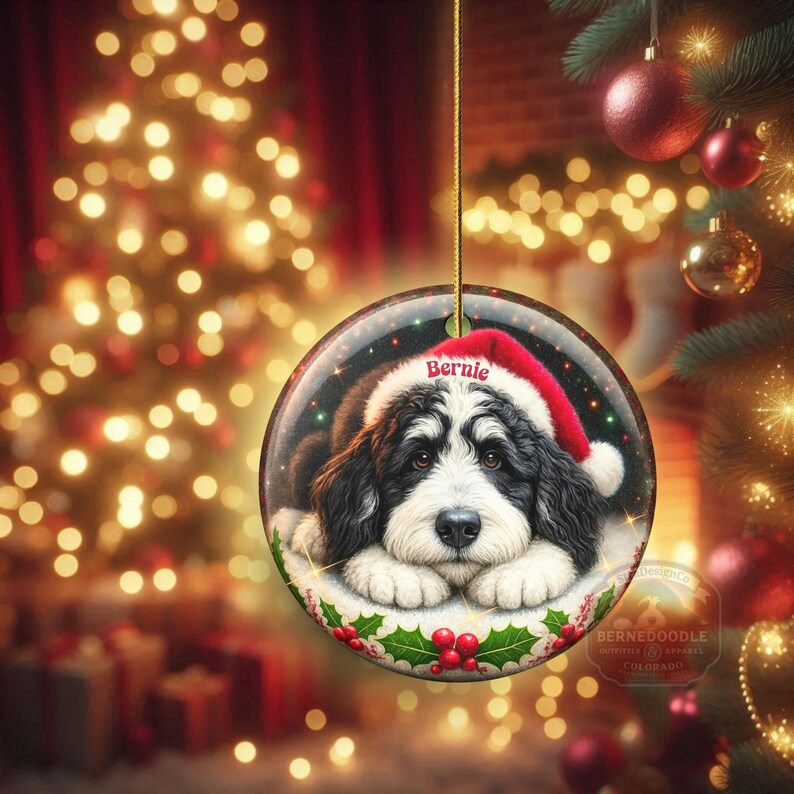 May include: Round Christmas ornament featuring a black and white Bernedoodle dog wearing a Santa hat with the name "Bernie". The ornament is decorated with holly and berries, and is hanging from a gold string. The background is a blurred Christmas scene.