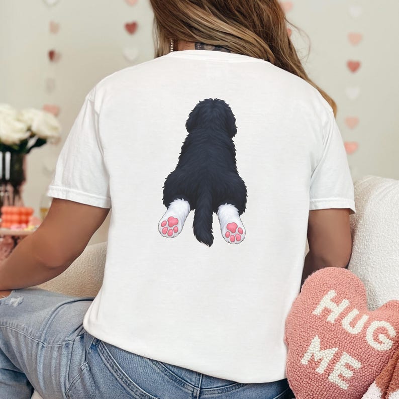 May include: White t-shirt featuring a graphic of a black and white dog's rear end with pink paw pads. The dog graphic is centered on the back of the shirt. The shirt is made of a soft material.