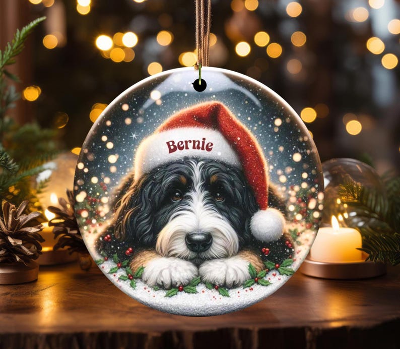 May include: A round Christmas ornament featuring a Bernese Mountain Dog wearing a Santa hat with the name "Bernie". The ornament has a snowy background with holly and berries around the dog's face. A festive holiday decoration.