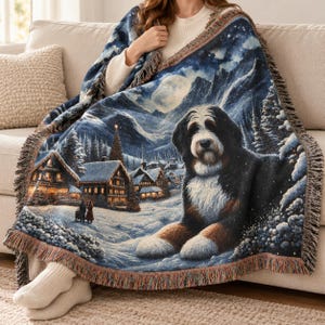 Puede incluir: Una manta tejida que presenta a un gran perro de monta&ntilde;a de Berna en primer plano, sobre un paisaje de pueblo invernal nevado con caba&ntilde;as iluminadas, pinos, una luna llena y monta&ntilde;as, con un borde de flecos decorativo.