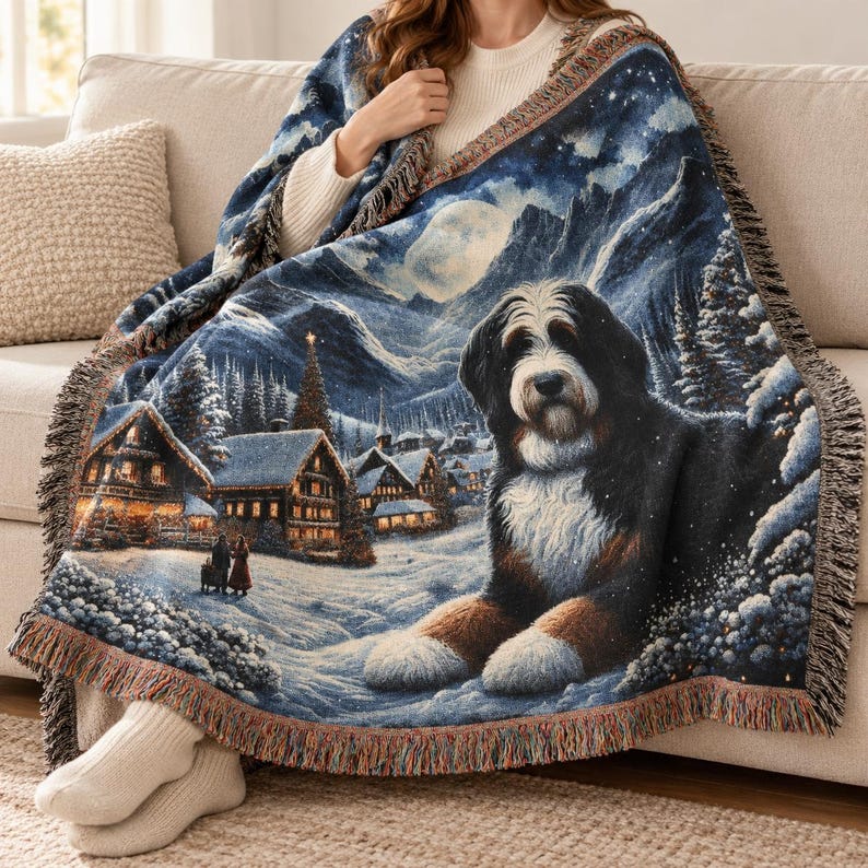 Puede incluir: Una manta tejida que presenta a un gran perro de monta&ntilde;a de Berna en primer plano, sobre un paisaje de pueblo invernal nevado con caba&ntilde;as iluminadas, pinos, una luna llena y monta&ntilde;as, con un borde de flecos decorativo.