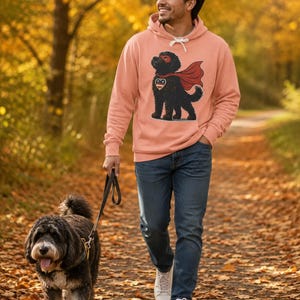 Pu&ograve; includere: Una felpa con cappuccio color pesca che presenta la grafica di un cane nero che indossa un mantello e una maschera da supereroe rossi, con un emblema a forma di cuore sul petto. La felpa ha maniche lunghe e una tasca a marsupio.