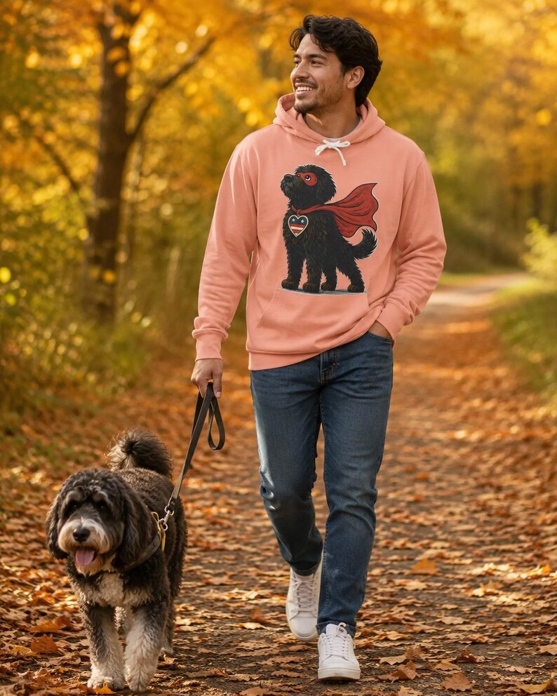 Pu&ograve; includere: Una felpa con cappuccio color pesca che presenta la grafica di un cane nero che indossa un mantello e una maschera da supereroe rossi, con un emblema a forma di cuore sul petto. La felpa ha maniche lunghe e una tasca a marsupio.