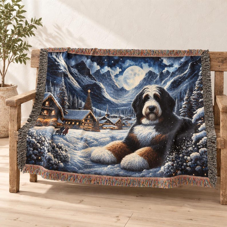 Puede incluir: Una manta tejida que presenta una escena detallada de un perro de monta&ntilde;a de Berna descansando en un paisaje nevado por la noche. La imagen incluye una luna llena, monta&ntilde;as nevadas, pinos, caba&ntilde;as iluminadas y bordes con flecos.
