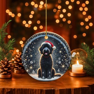 May include: Round Christmas ornament featuring a black dog wearing a Santa hat and red collar. The ornament has a snowy background with twinkling lights and snowflakes. The dog's name, "Teddy", is on the hat. A gold string is attached for hanging.