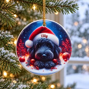 May include: A round ceramic Christmas ornament featuring a black dog wearing a red and white Santa hat. The name Tilly is printed in red on the hat brim. The background shows red berries and ornaments against a dark blue snowy night sky.