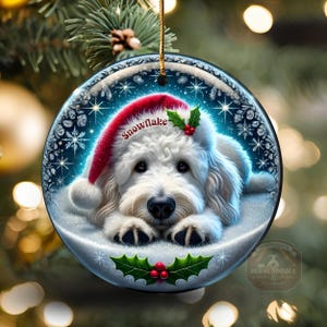 May include: A round Christmas ornament featuring a white dog wearing a red Santa hat with the word "Snowflake". The ornament has a winter scene with snowflakes and holly. A gold string is attached for hanging.