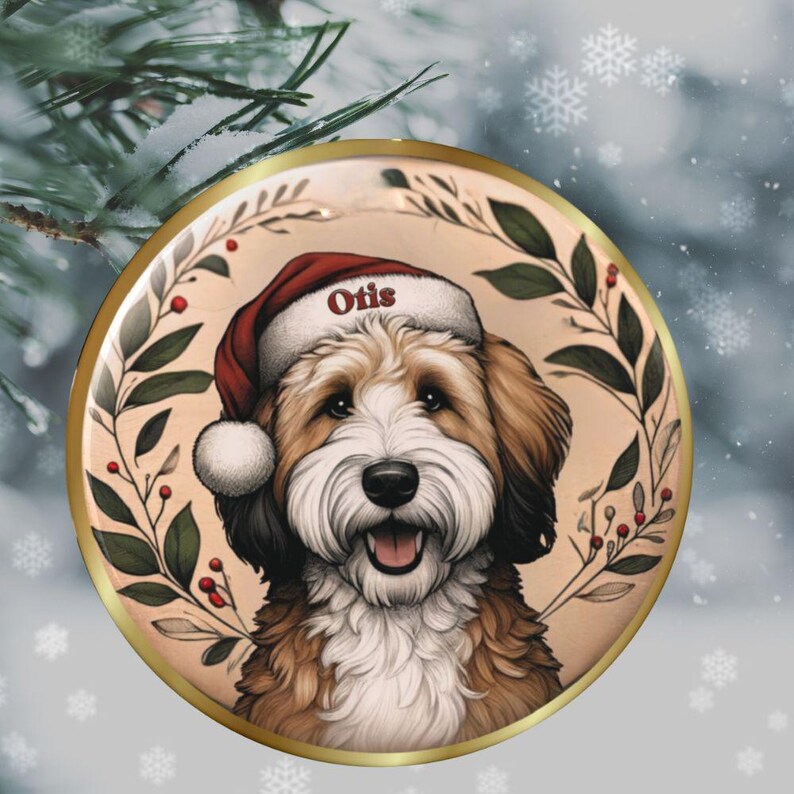 Puede incluir: Un adorno navide&ntilde;o redondo con un perro marr&oacute;n y blanco que lleva un gorro de Pap&aacute; Noel rojo con el nombre Otis impreso en el borde. El perro est&aacute; enmarcado por una corona circular de hojas verdes y bayas rojas sobre un fondo beige.