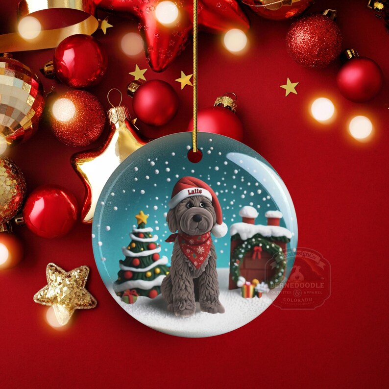 May include: A round Christmas ornament featuring a brown dog wearing a Santa hat and a red bandana. The ornament has a snowy scene with a Christmas tree and a decorated house. The background is red with Christmas decorations.
