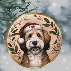 May include: A round Christmas ornament featuring a smiling dog wearing a Santa hat with the name "Otis". The dog is surrounded by a wreath of green leaves and red berries, with a gold border. The background has a snowy effect.