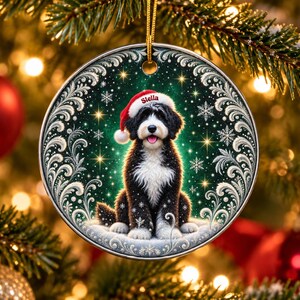 May include: A round ceramic Christmas ornament featuring a black and white Bernedoodle dog wearing a red Santa hat with the name Stella printed on the brim. The dog sits against a dark green background with white snowflakes and swirling filigree.