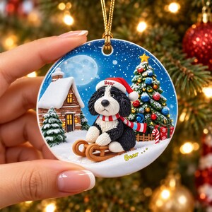 A hand holding a round ornament featuring a black and white dog wearing a Santa hat and scarf, sitting on a sled in front of a snowy village scene with a Christmas tree and house.
