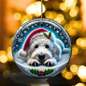 May include: A round Christmas ornament featuring a white dog wearing a red Santa hat with the word "Snowflake". The ornament has a snowy background with holly and berries, and a gold string for hanging.
