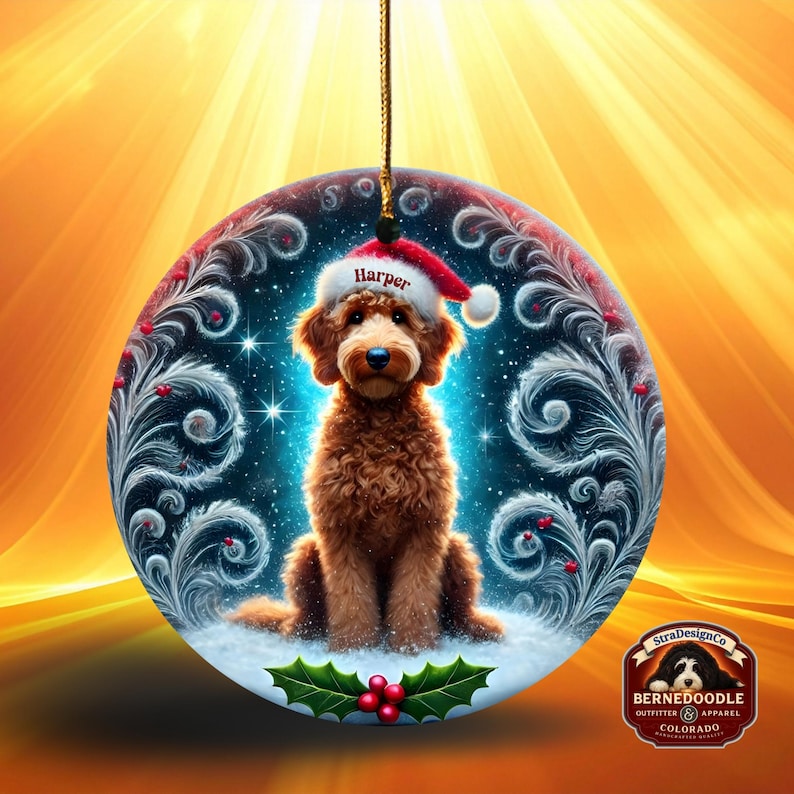A round ceramic Christmas ornament showing a curly brown goldendoodle dog seated in snow, wearing a red Santa hat with white trim. The background glows with blue light, swirling frosted feather details, winter stars, and holly leaves with red berries near the bottom.