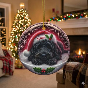 May include: Round Christmas ornament featuring a black dog wearing a Santa hat with the name "Teddi". The ornament has a red and white design with snowflakes and holly accents. A gold string is attached for hanging.