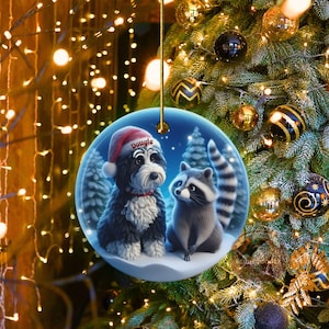 A 3-inch double-sided ceramic Christmas ornament featuring an adorable claymation-style Bernedoodle wearing a red Santa hat personalized  sitting beside a cute raccoon under a snowy winter night sky. The ornament has crisp detailing, soft blue tones, and a warm Christmas glow in the blurred background. Perfect for Bernedoodle lovers, handmade Christmas gift shoppers. High-quality ceramic with vibrant colors, ideal for tree decorating, pet memorial ornaments, or festive seasonal displays.