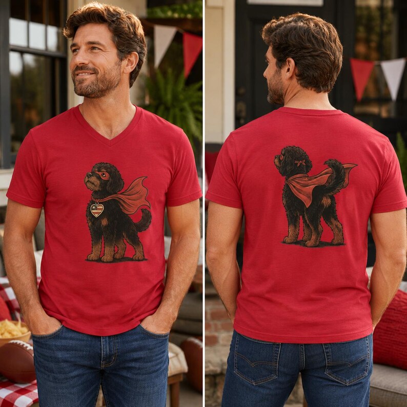 May include: A red V-neck t-shirt featuring a graphic of a black dog wearing a red superhero cape, a red eye mask, and a heart-shaped pendant with a flag pattern. The back of the shirt displays the same black dog in a superhero cape from behind.