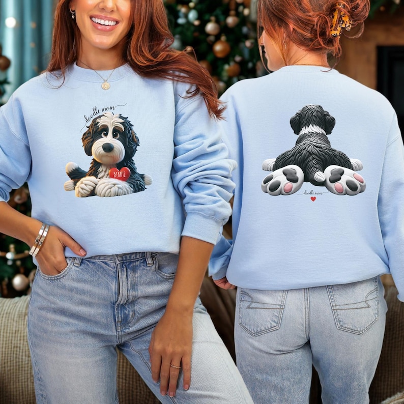 A woman wearing a Bernedoodle sweatshirt with cute design on the front and the back.  The front features a bernedoodle dog in claymation design holding a heart with his name, across the top of his head the words, "Doodle Mom", on the back, it shows the bernedoodle laying rear view with the pads of his doodle feet flipped back in a cute pose, with a heart and the words, "Doodle Mom".