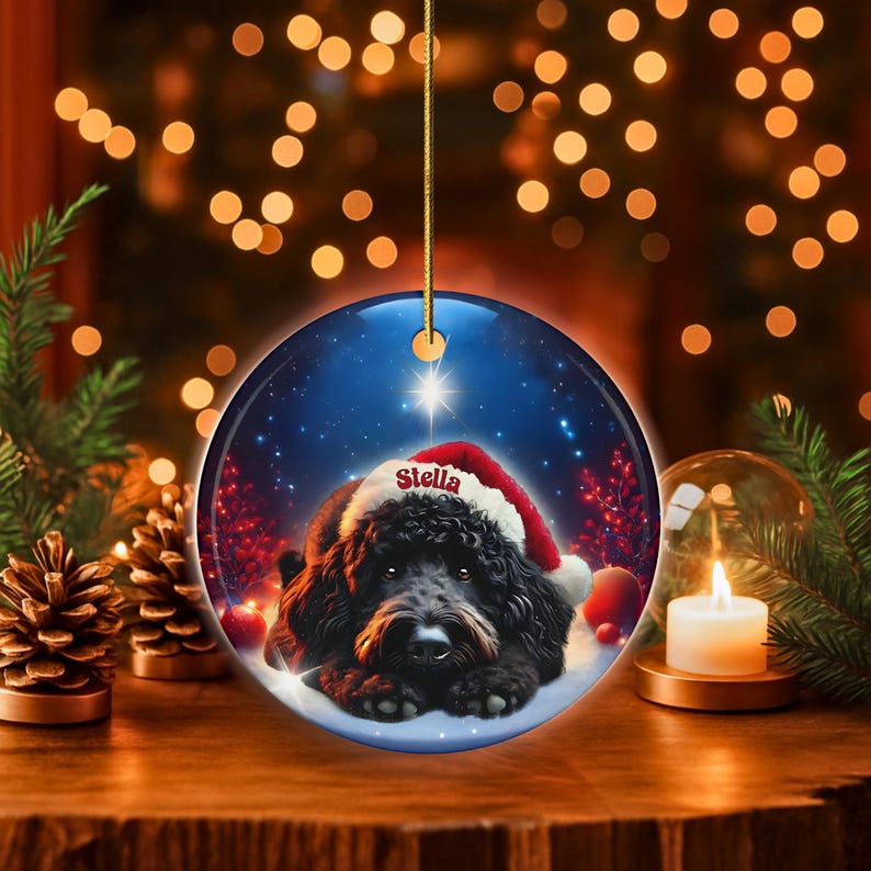 May include: Round Christmas ornament featuring a black dog wearing a Santa hat, with the name "Stella" on the hat. The ornament has a blue background with a star and snow. A gold string is attached for hanging.