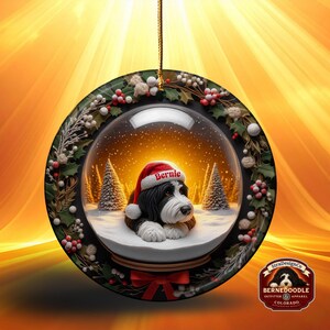 May include: A round Christmas ornament featuring a snow globe design. Inside the globe is a black and white dog wearing a Santa hat with the name "Bernie". The ornament is decorated with holly and berries, and has a red bow at the bottom.