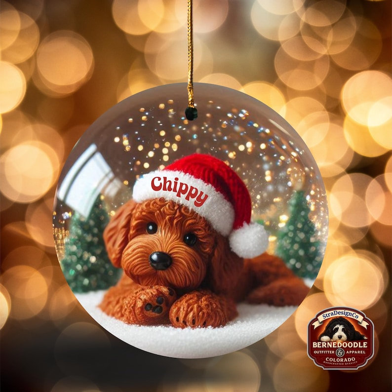 May include: A clear glass Christmas bauble featuring a brown dog wearing a red Santa hat with the name "Chippy". The bauble has a gold string for hanging and includes a small logo.