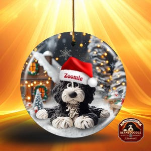 May include: A round Christmas ornament featuring a black and white Bernedoodle dog wearing a red Santa hat with the name "Zoomie". The ornament has a winter scene background with a house and trees. A gold string is attached for hanging.