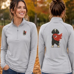 K&ouml;nnte beinhalten: Ein hellgrauer Langarm-Pullover mit kurzem Rei&szlig;verschluss, der auf der linken Brust ein kleines Motiv eines Berner Sennenhundes mit rotem Umhang und auf der R&uuml;ckseite ein gr&ouml;&szlig;eres, passendes Motiv des Hundes mit einer herzf&ouml;rmigen US-Flagge zeigt.