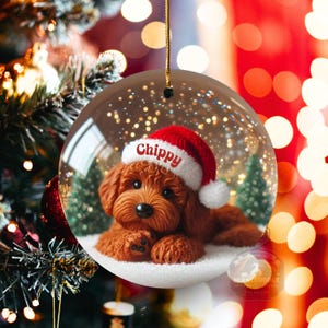May include: A clear, round Christmas bauble featuring a brown dog wearing a red Santa hat with the name "Chippy" on it. The dog is lying on a bed of white snow with small green trees in the background. The bauble is suspended from a gold string.