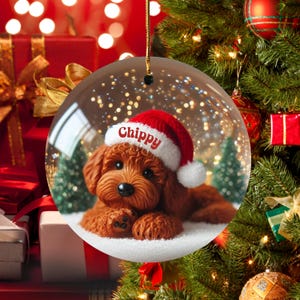 May include: A clear, round Christmas bauble featuring a brown dog wearing a red Santa hat with the name "Chippy" in white. The dog is lying on a bed of white snow, with small green trees in the background. The bauble hangs from a gold string.