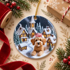 May include: A round ceramic Christmas ornament featuring a golden-brown curly-haired dog wearing a white Santa hat with the name Maggie printed in red. The background depicts a snowy village scene with houses and a church at night.
