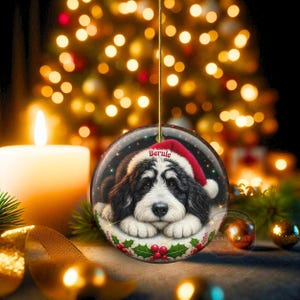 May include: A round Christmas ornament featuring a black and white dog wearing a Santa hat with the name "Bernie". The ornament is hanging from a gold string, with a blurred background of Christmas lights and a lit candle.