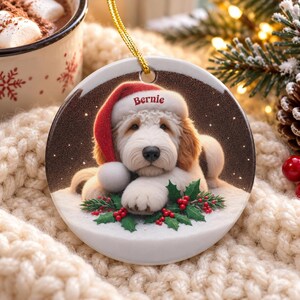 May include: A round ceramic Christmas ornament featuring a fluffy white and tan dog wearing a red Santa hat with the name Bernie printed on the brim. The dog rests on a snowy surface decorated with green holly leaves and red berries.