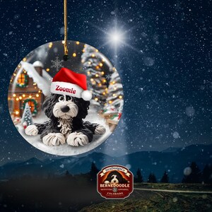 May include: A round Christmas ornament featuring a black and white Bernedoodle dog wearing a red Santa hat with the name "Zoomie". The ornament has a winter scene background with a snowy landscape and a house with Christmas lights. The ornament is hanging by a gold string.