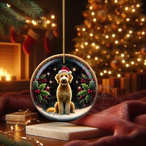May include: A round Christmas ornament featuring a golden doodle dog wearing a Santa hat. The ornament has a snowy scene with holly and berries. The background includes a Christmas tree and a fireplace.