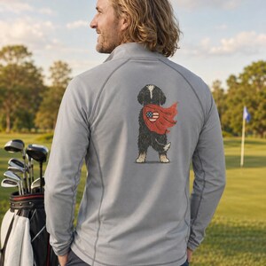 K&ouml;nnte beinhalten: Ein hellgrauer Langarm-Pullover mit kurzem Rei&szlig;verschluss, der auf der R&uuml;ckseite eine Grafik eines schwarz-wei&szlig;en Hundes zeigt, der einen roten Umhang mit einem herzf&ouml;rmigen amerikanischen Flaggendesign tr&auml;gt. Das Kleidungsst&uuml;ck hat Raglan&auml;rmel.