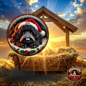 May include: Round Christmas ornament featuring a black dog wearing a Santa hat, with the name "Stella" on the hat. The ornament has a festive design with holly and berries. The background shows a wooden structure and hay.