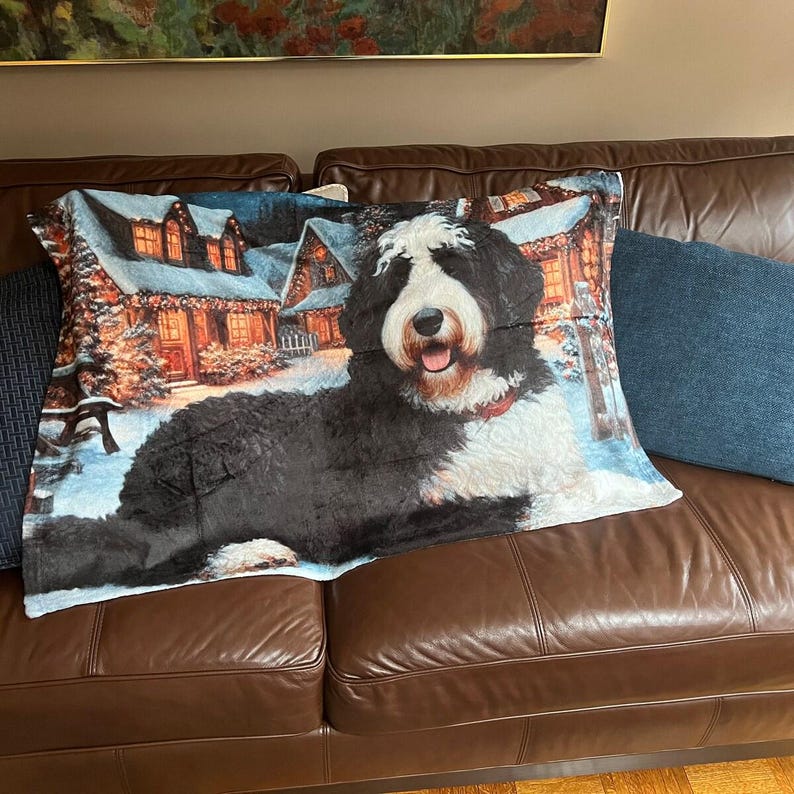 May include: A decorative throw pillow featuring a Bernedoodle dog with a red collar, set against a winter scene with snow-covered houses and trees. The dog is black, white, and brown, resting on a brown leather couch.